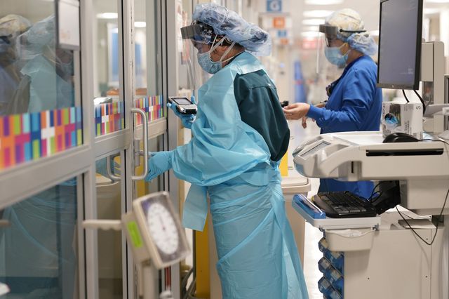 Medical workers wear gowns, masks, gloves, hair coverings, and a face shield in a hospital corridor